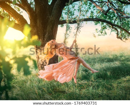 Beautiful happy woman nymph sitting on swings. Magical fantasy swing. princess long peach color orange silk vintage dress fluttering wind. blond hair fly in motion. Tree sunshine green grass forest