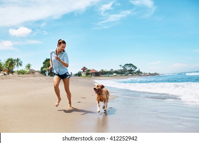 Beautiful Happy Woman Running With Her Dog, Golden Retriever On Wet Sand On The Beach By Sea. Girl Enjoying Summer Holidays Vacations, Having Fun With Her Pet. Summertime Concept.