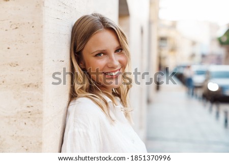 Beautiful happy woman smiling and looking at camera while leaning on wall at city street