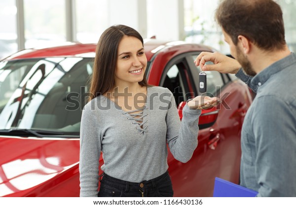 Beautiful and happy\
woman smiling, looking at man and receiving keys from her new red\
car. Male manager selling automobile in car dealership to female\
customer and giving her\
keys.