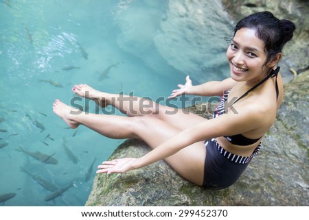 Beautiful happy woman in swimsuit showing fish in clear water. Asian girl sitting on a rock above a tropical pond full of fish. 