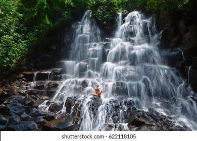 Beautiful happy woman travel in Bali jungle. Sit in zen-like yoga pose under flowing spring water, enjoy tropical cascade waterfall. Nature day tour, walking adventure, fun on family summer vacation.