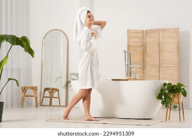Beautiful happy woman wearing white robe near tub in bathroom