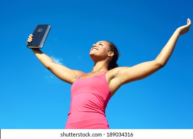 Beautiful happy young african american female celebrating success with a Bible against cloudy sky. With arms up praising God. Horizontal Shot.