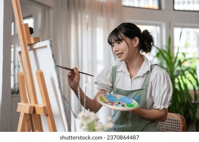 A beautiful and happy young Asian female artist is enjoying painting on a canvas in her home studio while working on her art project. creativity hobby concept - Powered by Shutterstock