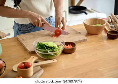 A beautiful, happy young Asian woman cutting a tomato on a cutting board, preparing ingredients for her healthy salad, enjoying cooking in the kitchen. home cooking, domestic life, healthy eating - Powered by Shutterstock