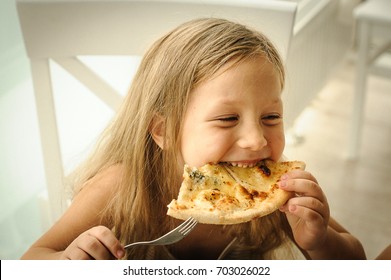 Beautiful happy young girl laugh and biting off big slice of fresh made pizza. She sit at white chair in Provence style interior, smile and enjoy sunny day and yummy meal. She has long blonde hair. 