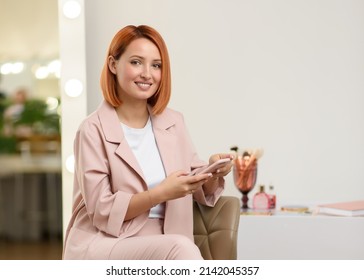 Beautiful Happy Young Red-haired Woman In A Casual Jacket Sits On A Chair In A Beauty Salon And Looks At The Camera. Attractive Smiling Caucasian Girl With A Bob Haircut Holds A Smartphone In Her Hand