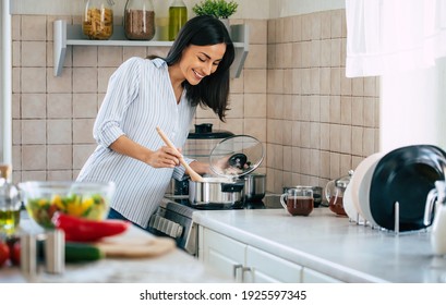 Beautiful happy young woman is cooking in the home kitchen and testing some soup from the pan on the stove - Powered by Shutterstock