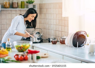 Beautiful happy young woman is cooking in the home kitchen and testing some soup from the pan on the stove