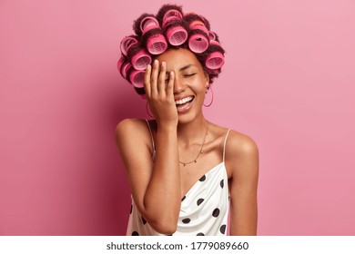 Beautiful happy young woman does hairstyle with hair rollers, makes face palm and cannot stop laughing, hears something very funny, dressed casually, spends free time at home. Hair care concept - Powered by Shutterstock