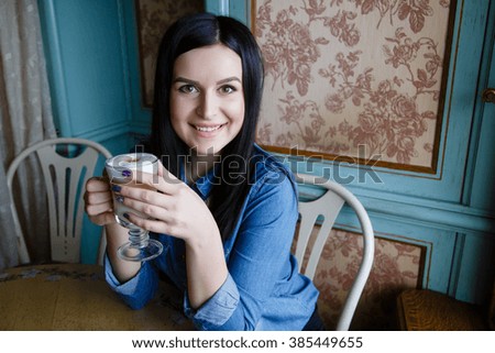 Beautiful happy young woman dressed in denim shirt sitting in the cafe and drinking coffee