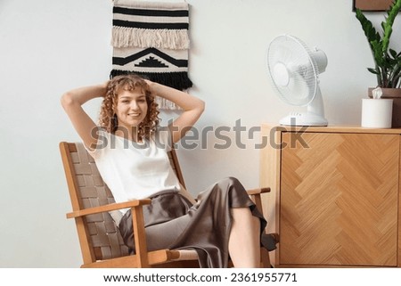 Beautiful happy young woman with electric fan sitting on chair at home