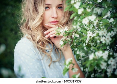 Beautiful happy young woman enjoying smell in a flowering spring garden. Blonde with blue eyes and blue dress.