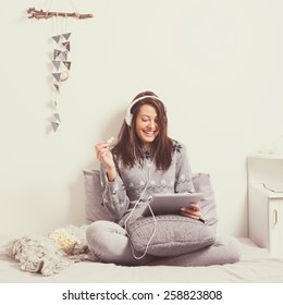 Beautiful happy young woman with headphones and tablet watching a movie on tablet in bed eating popcorn. Mild retouch, matte color filter, contemporary look. - Powered by Shutterstock