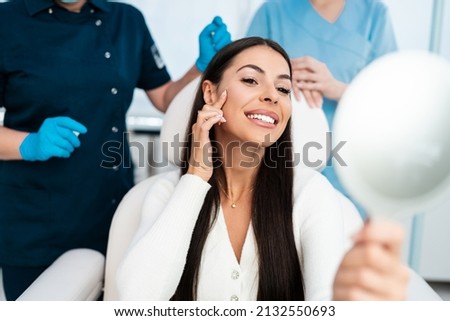 Beautiful and happy young woman sitting in medical chair and looking in the mirror. She is satisfied after successful beauty treatment with hyaluronic acid fillers or botulinum toxin injections.