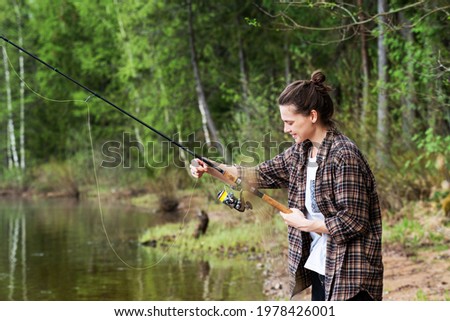 Beautiful happy young woman standing on the shore of the lake with a fishing rod in her hands, summer vacation 