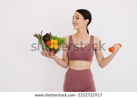 Beautiful happy young woman with tray of various healthy vegetables and showing her muscles with dumbbell isolated on the white background Stok fotoğraf © 
