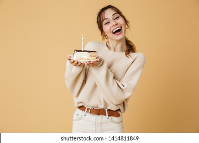 Beautiful happy young woman wearing sweater standing isolated over beige background, holding birthday cake with candle on a plate
