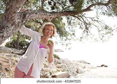 Beautiful Healthy Senior Woman Relaxing On Coastal Destination, Being Playful Around Tree Trunk, Eating A Green Apple On Holiday, Nature Outdoors. Healthy Eating Lifestyle, Mature Woman, Sea Exterior.