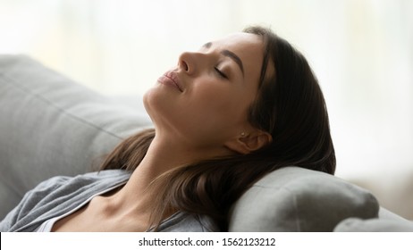Beautiful healthy young woman taking deep breath of fresh air relaxing napping on comfortable couch, attractive calm lady meditating enjoying resting on sofa feel peace of mind at home, close up view
