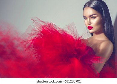 Beautiful High fashion woman in red dress posing in studio. Gorgeous brunette model girl with beautiful hairstyle and make-up, Wearing Stunning evening red dress with long gown of tulle and smoke