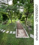 A beautiful high-angle view of a stone paved pathway or staircase leading downwards through a vibrant green park. The walkway is lined with small decorative plants, lush lawns, stepping stones on the side, and shady tropical trees, creating a peaceful, relaxing, and refreshing natural atmosphere