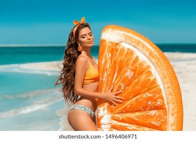 Beautiful hippie girl portrait with orange earring by Inflatable pool float mattress on tropical beach. Fit Brunette with long healthy curly hair and makeup on summer vacation.
