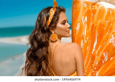 Beautiful Hippie Girl Portrait With Orange Earring By Inflatable Pool Float Mattress On Tropical Beach. Brunette With Long Healthy Curly Hair And Makeup On Summer Vacation.