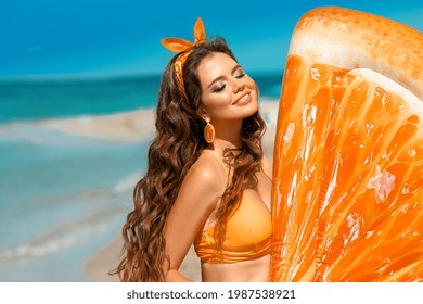 Wunderschönes Hippie-Girl-Portrait mit Orangenohrring von Aufblasbaren Schwimmbadmatratze am tropischen Strand. Brunette mit langen gesunden, lockigen Haaren und Make-up im Sommerurlaub.