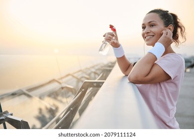 Beautiful Hispanic Athlete Woman In Pink T-shirt And Terry Wristbands Smiles Toothy Smile Standing On City Bridge With Bottle Of Water And Puts On Earphones, Resting After Early Morning Jog At Sunrise