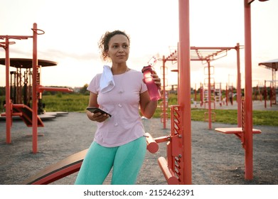 Beautiful Hispanic Athletic Woman Resting On A Sports Field After Workout On Stationary Gym Machines And Crossbar, Holding A Bottle With Water And Smartphone And Looking Away Against Sunset Background