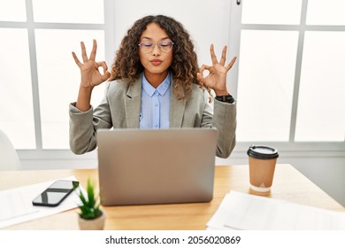 Beautiful Hispanic Business Woman Sitting On Desk At Office Working With Laptop Relax And Smiling With Eyes Closed Doing Meditation Gesture With Fingers. Yoga Concept. 