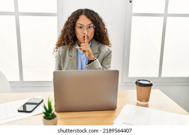 Beautiful hispanic business woman sitting on desk at office working with laptop asking to be quiet with finger on lips. silence and secret concept. 