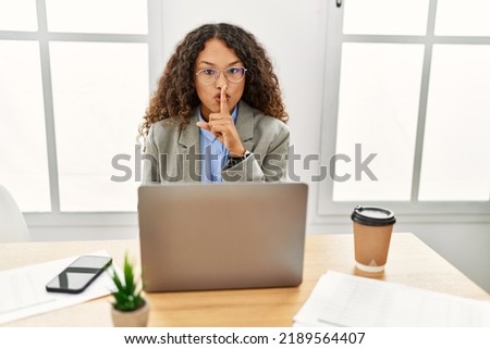 Beautiful hispanic business woman sitting on desk at office working with laptop asking to be quiet with finger on lips. silence and secret concept. 