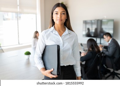 Beautiful hispanic businesswoman holding a folder standing in office board room with colleagues meeting in background