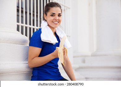 Beautiful Hispanic Female Athlete Taking A Break From Running And Eating A Banana