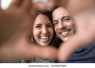 Beautiful Hispanic wife and husband look at camera through fingers showing symbol of love, close up faces view. Romantic couple make selfie picture, celebrate anniversary, engagement or Valentines Day - Powered by Shutterstock