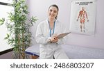 Beautiful hispanic woman in a clinic room with clipboard, wearing a lab coat and a stethoscope smiling at the camera
