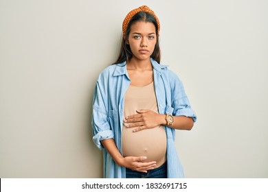 Beautiful Hispanic Woman Expecting A Baby, Touching Pregnant Belly Relaxed With Serious Expression On Face. Simple And Natural Looking At The Camera. 