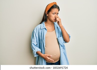 Beautiful Hispanic Woman Expecting A Baby, Touching Pregnant Belly Smelling Something Stinky And Disgusting, Intolerable Smell, Holding Breath With Fingers On Nose. Bad Smell 