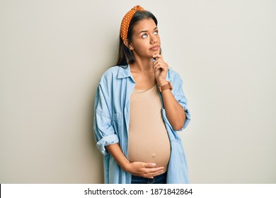 Beautiful Hispanic Woman Expecting A Baby, Touching Pregnant Belly With Hand On Chin Thinking About Question, Pensive Expression. Smiling With Thoughtful Face. Doubt Concept. 