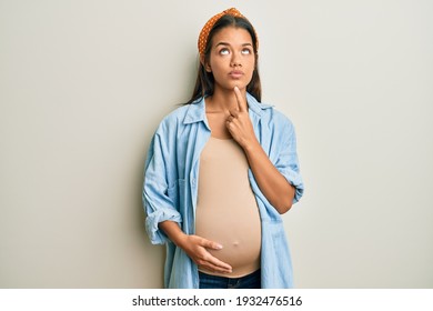 Beautiful Hispanic Woman Expecting A Baby, Touching Pregnant Belly Thinking Concentrated About Doubt With Finger On Chin And Looking Up Wondering 