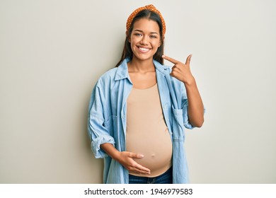 Beautiful Hispanic Woman Expecting A Baby, Touching Pregnant Belly Smiling Cheerful Showing And Pointing With Fingers Teeth And Mouth. Dental Health Concept. 