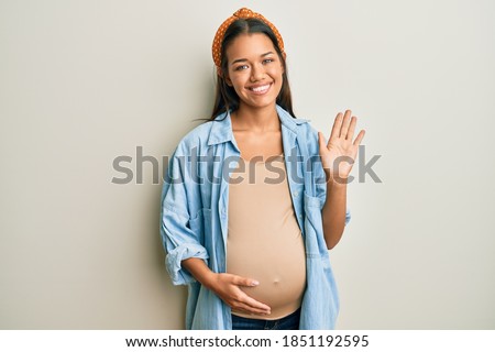 Beautiful hispanic woman expecting a baby, touching pregnant belly waiving saying hello happy and smiling, friendly welcome gesture 