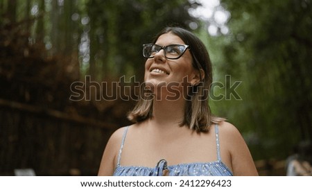 Beautiful hispanic woman with glasses, smiling confidently as she stands amongst kyoto's lush bamboo forest, looking around, full of joy and amazement.