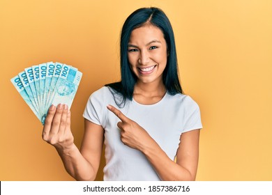Beautiful hispanic woman holding 100 brazilian real banknotes smiling happy pointing with hand and finger 