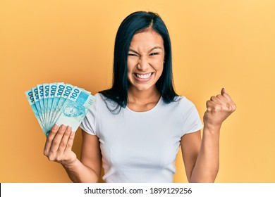 Beautiful hispanic woman holding 100 brazilian real banknotes screaming proud, celebrating victory and success very excited with raised arm 