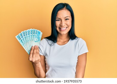 Beautiful hispanic woman holding 100 brazilian real banknotes looking positive and happy standing and smiling with a confident smile showing teeth 