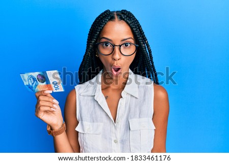 Beautiful hispanic woman holding 100 swiss franc banknote scared and amazed with open mouth for surprise, disbelief face 